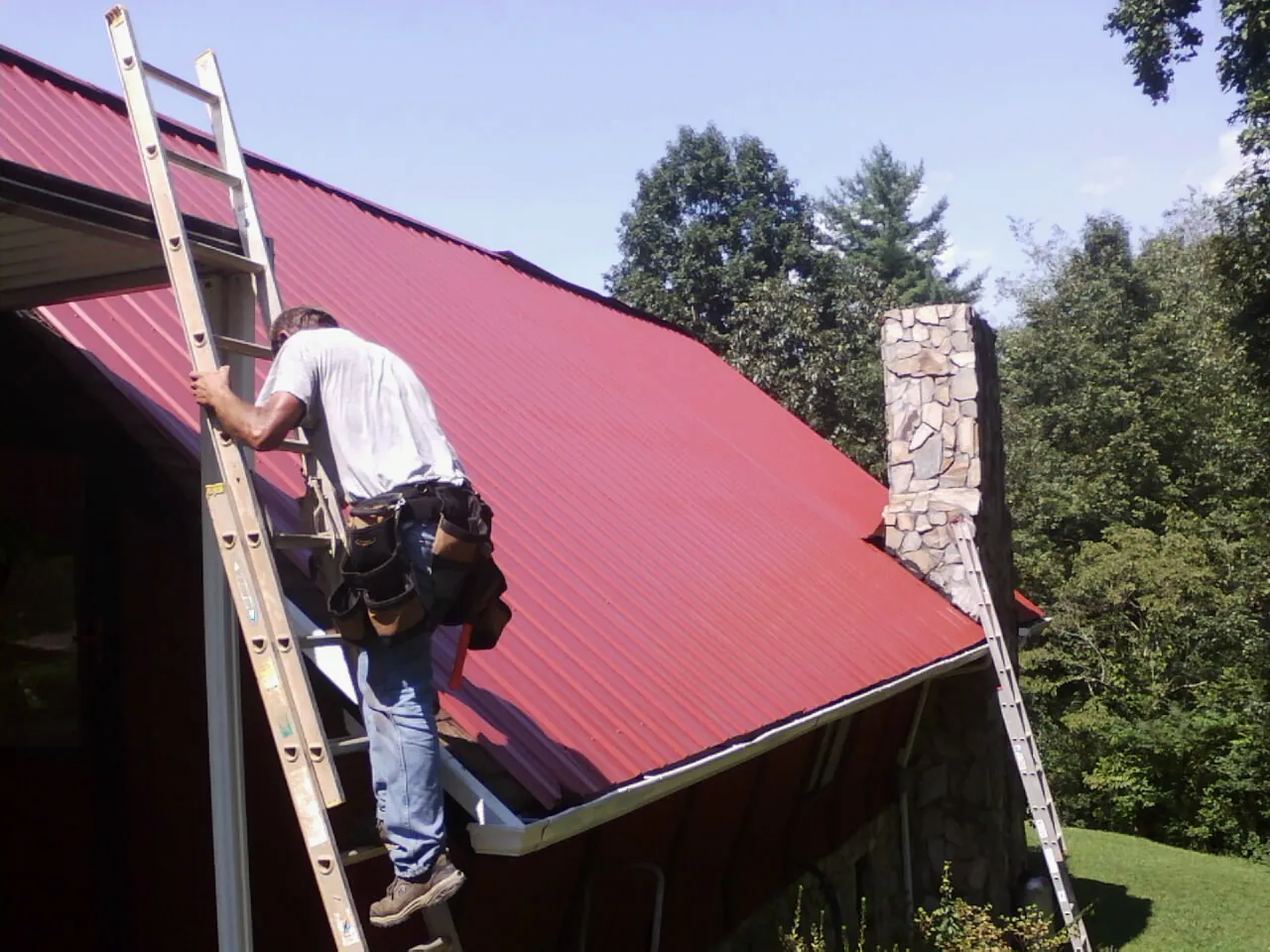 Roofer on a ladder beside a red metal roof and stone chimney — Soffit & Fascia Repair by Roofing Brothers in Drewsey
